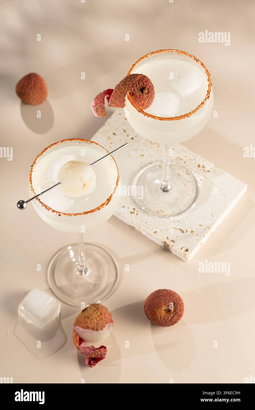 Oberblick auf zwei gekühlte Litschi-Cocktails in Stielgläsern mit Eiswürfeln, Obstgarnitur und pfeffrigem Zuckerrand, arrangiert mit frischer Litschi und be Stockfoto