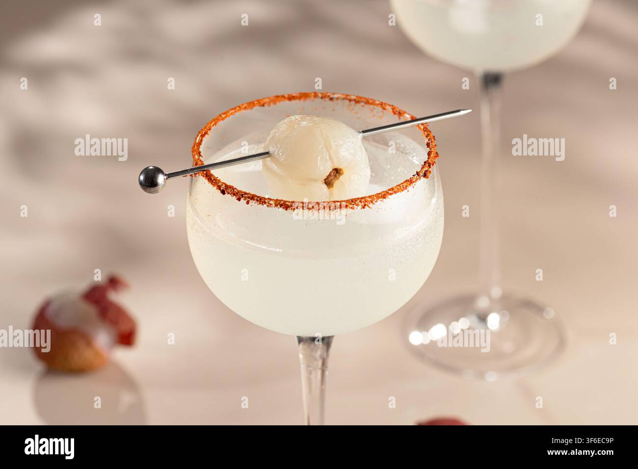 Ge kühlter Litschi-Cocktail in einem Stielglas mit rotem Gewürzrand, aufgespießter Litschigarne und frischer Litschifrucht auf einem weichen neutralen Hintergrund. Stockfoto