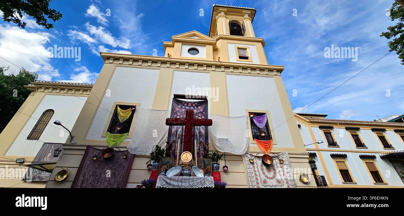 Rotes florales Kreuz, das vor einer mit Textilien, Keramikobjekten und religiösen Bildern geschmückten Kirchenfassade während des Cruces de Mayo-Festivals ausgestellt ist Stockfoto