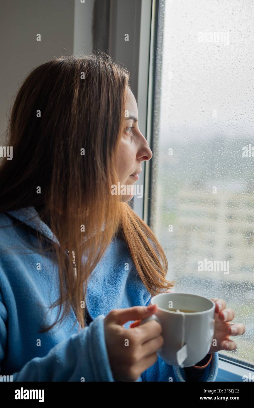 Frau schaut aus einem nassen Fenster mit Regentropfen, während sie eine warme Tasse Tee hält Stockfoto