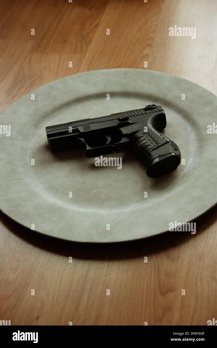 Schwarze Handfeuerwaffe ruht auf einem großen Keramikteller auf einem Holztisch und schafft ein surreales Konzept von Waffe und Gewalt Stockfoto
