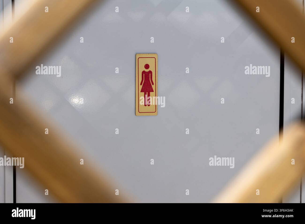 Ein Schild im Retro-Stil, das eine Damen-Toilette anzeigt, ist auf einer hellfarbenen Wand mit einem dezenten Rautenmuster zentriert, umrahmt von verschwommenen Holzelementen Stockfoto