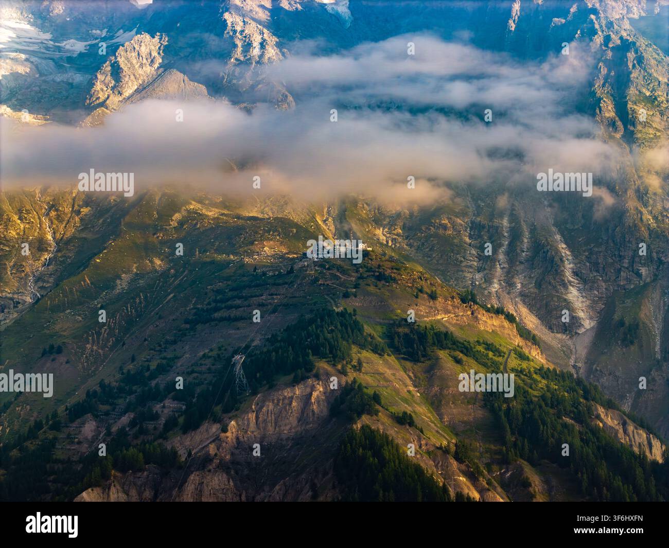 Luftbild des Eingangs zum Mont-Blanc-Tunnel, eingebettet zwischen den schroffen, wolkenbedeckten Gipfeln der Alpen, mit üppigen grünen Hängen und felsigem Gelände in C Stockfoto