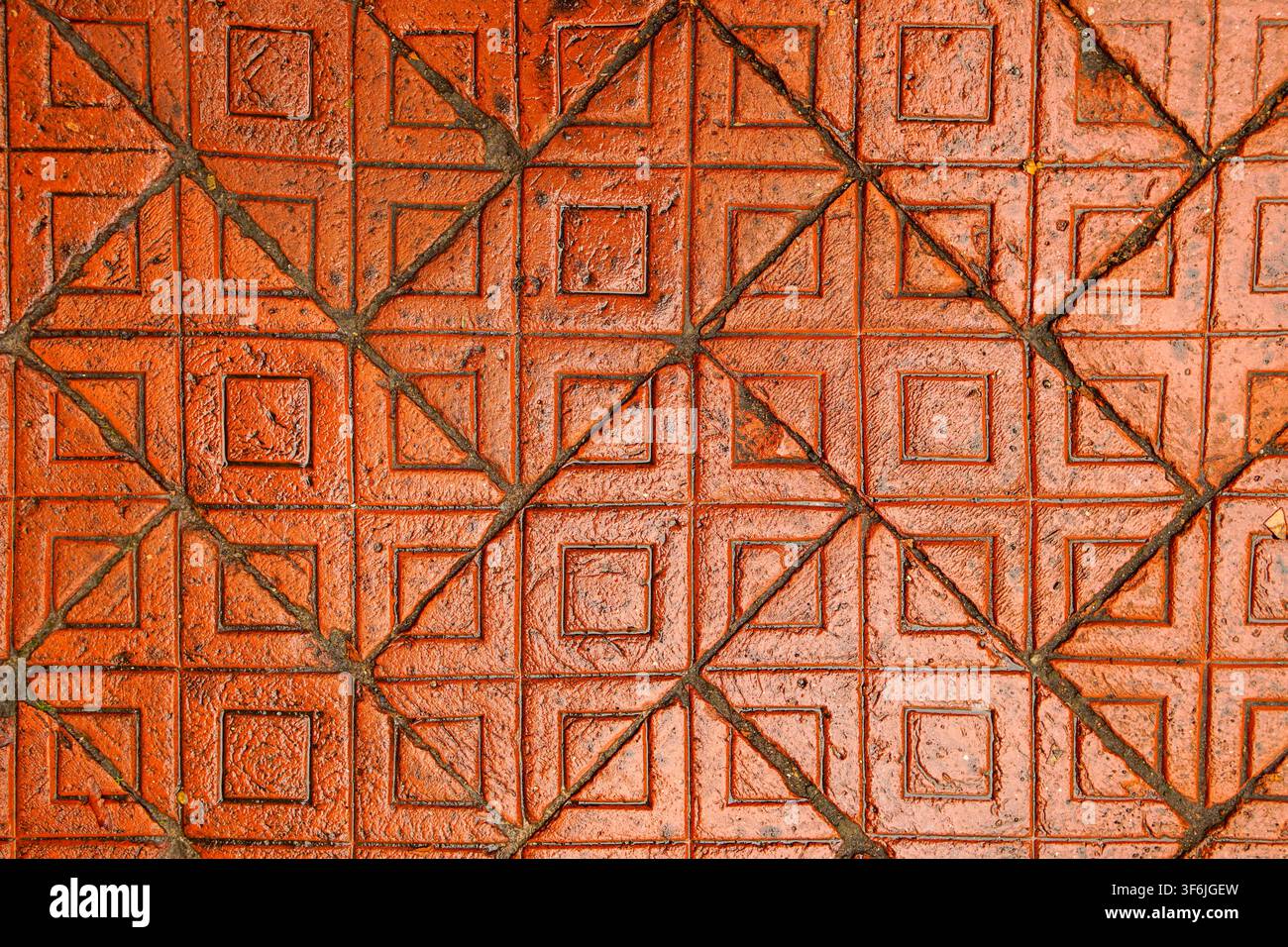 Nahaufnahme von oben auf verwitterte rot-orangefarbene Pflastersteine mit einem erhabenen quadratischen und diagonalen Kreuzmuster, getrennt durch dunkle gerillte Linien Stockfoto