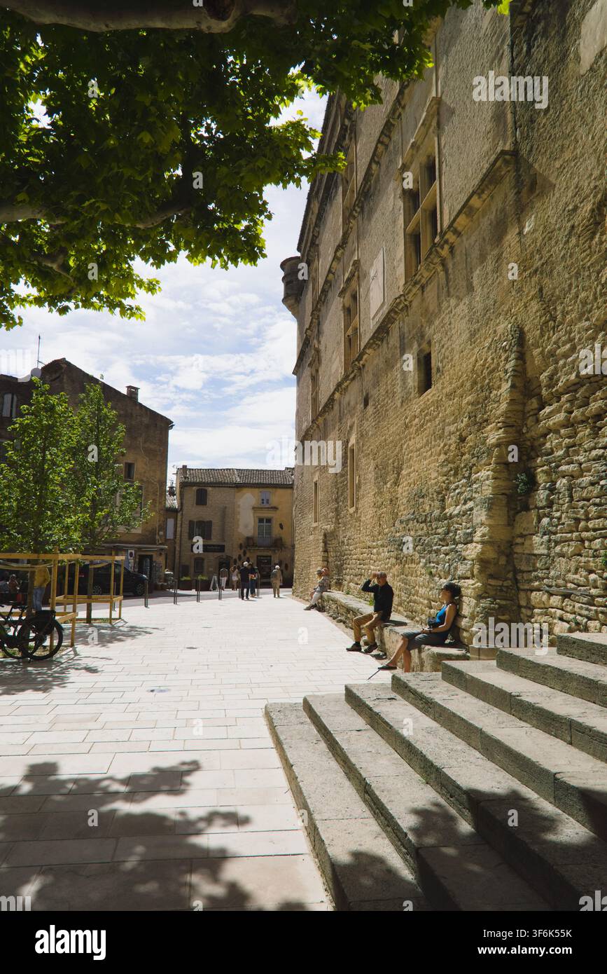 Touristen ruhen auf Steinbänken entlang der Burgmauer im sonnenbeschienenen Platz von Gordes, Provence, Frankreich. Stockfoto