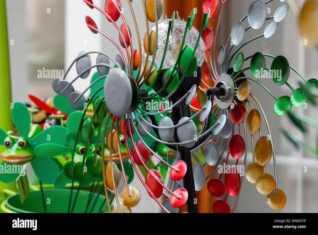 Dekorativer Windspiel mit metallenen Flügeln in verschiedenen Farben, umgeben von fröhlichen Gartenornamenten in einer lebhaften Außenumgebung Stockfoto