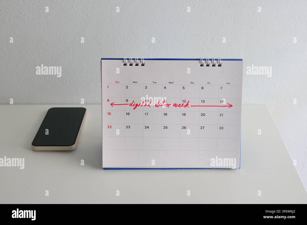 Selektive Fokussierung auf die handgeschriebene Phrase "digitale Entgiftungswoche" in roter Tinte auf dem Schreibtischkalender, daneben liegt ein ausgeschaltetes Smartphone. Das Arrangement befindet sich auf Stockfoto