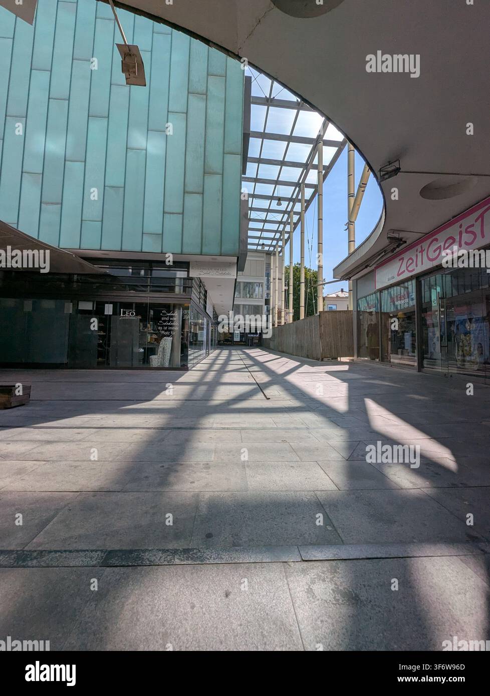 Sonnenbeschienene Fußgängerpassage in einer modernen Einkaufspassage mit gebogenen Glasdachplatten, hohen Säulen, blauen Ladenfronten, Steinpflaster und langen Schatten Stockfoto
