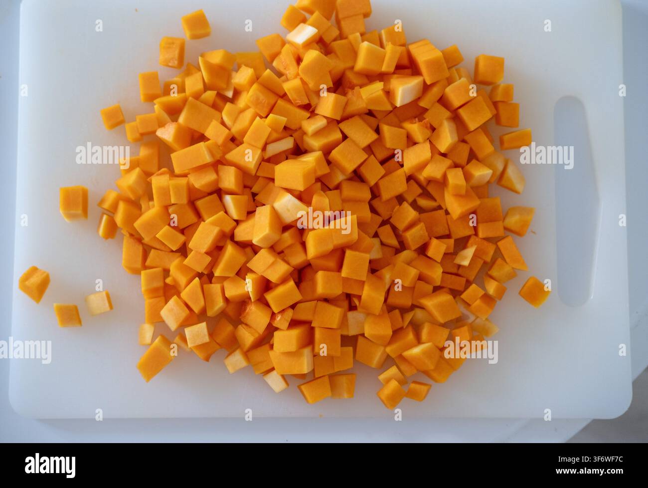 Ein Butternusskürbis wird auf einem weißen Kunststoffschneidebrett in kleine Würfel gewürfelt. Stockfoto