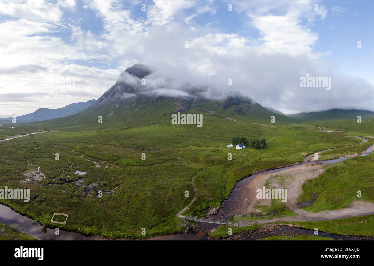Luftbild von Buchaille Etive Mor, einem Berg in Glen Coe, der teilweise von Wolken verhüllt ist, üppige grüne Hügel, ein gewundener Fluss und ein kleines weißes Haus in Stockfoto
