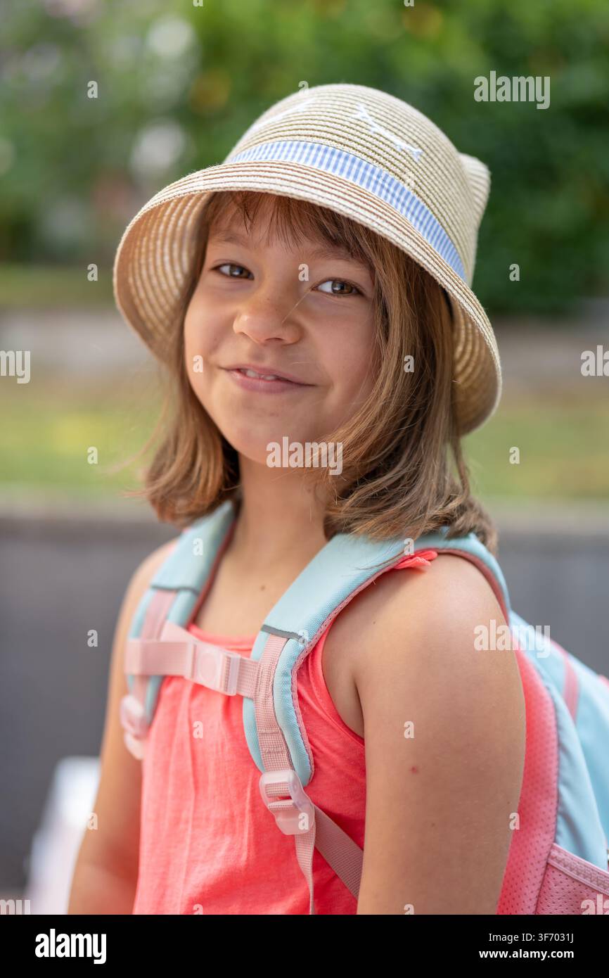 Lächelndes achtjähriges Mädchen trägt einen Sommerhut und steht im Freien mit einem Rucksack über den Schultern Stockfoto
