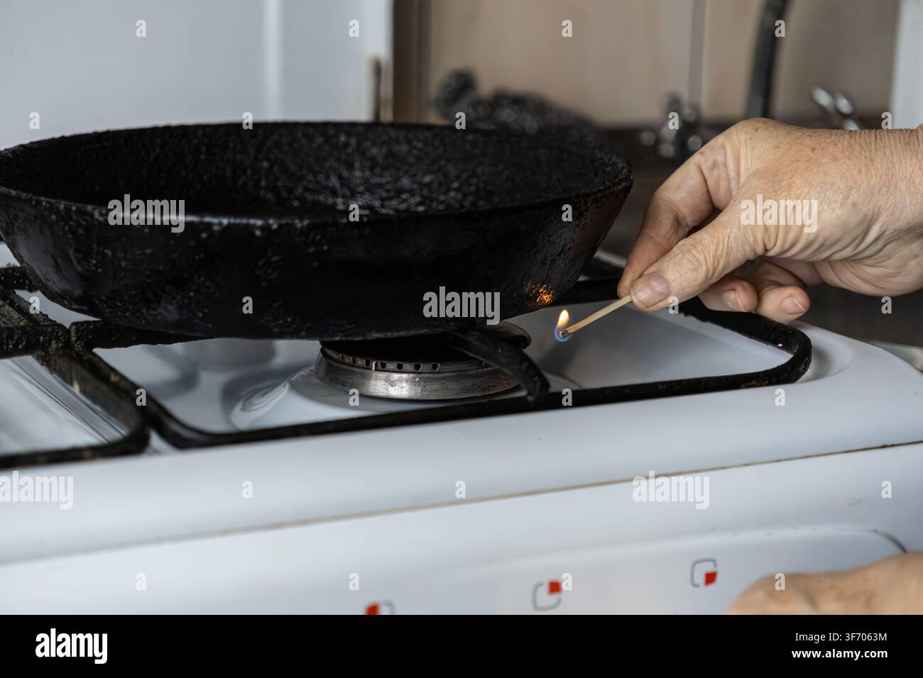 Reife Hand zündet einen Gasherd in der Küche mit einem kleinen Holzschuh an, um eine Vintage-Bratpfanne zu erwärmen, was die nostalgische Mahlzeitvorbereitung, Zuhause, repräsentiert Stockfoto