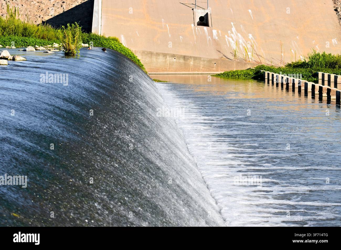 Wasser fließt über einen gebogenen Betonüberlauf in einen breiten Kanal, der von Hochwasserschutzbauwerken und grüner Vegetation umgeben ist. Stockfoto