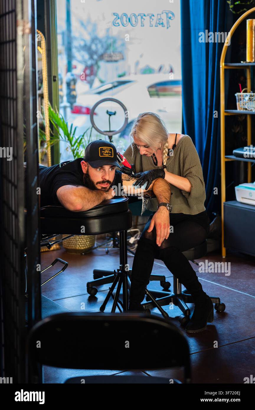 März 2023: Ein Tätowierer tätowiert den Arm eines Kunden in der Nähe der offenen Tür eines Studios, wobei die umgekehrte Ladenschilder in dem Fenster sichtbar ist. Stockfoto