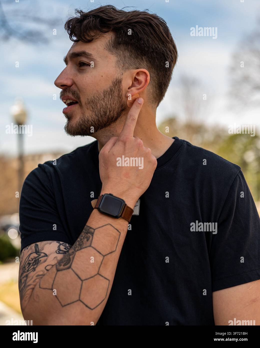 Ein junger bärtiger Mann in einem schwarzen T-Shirt wird im Profil im Freien fotografiert, berührt sein Ohr und schaut zur Seite. Stockfoto