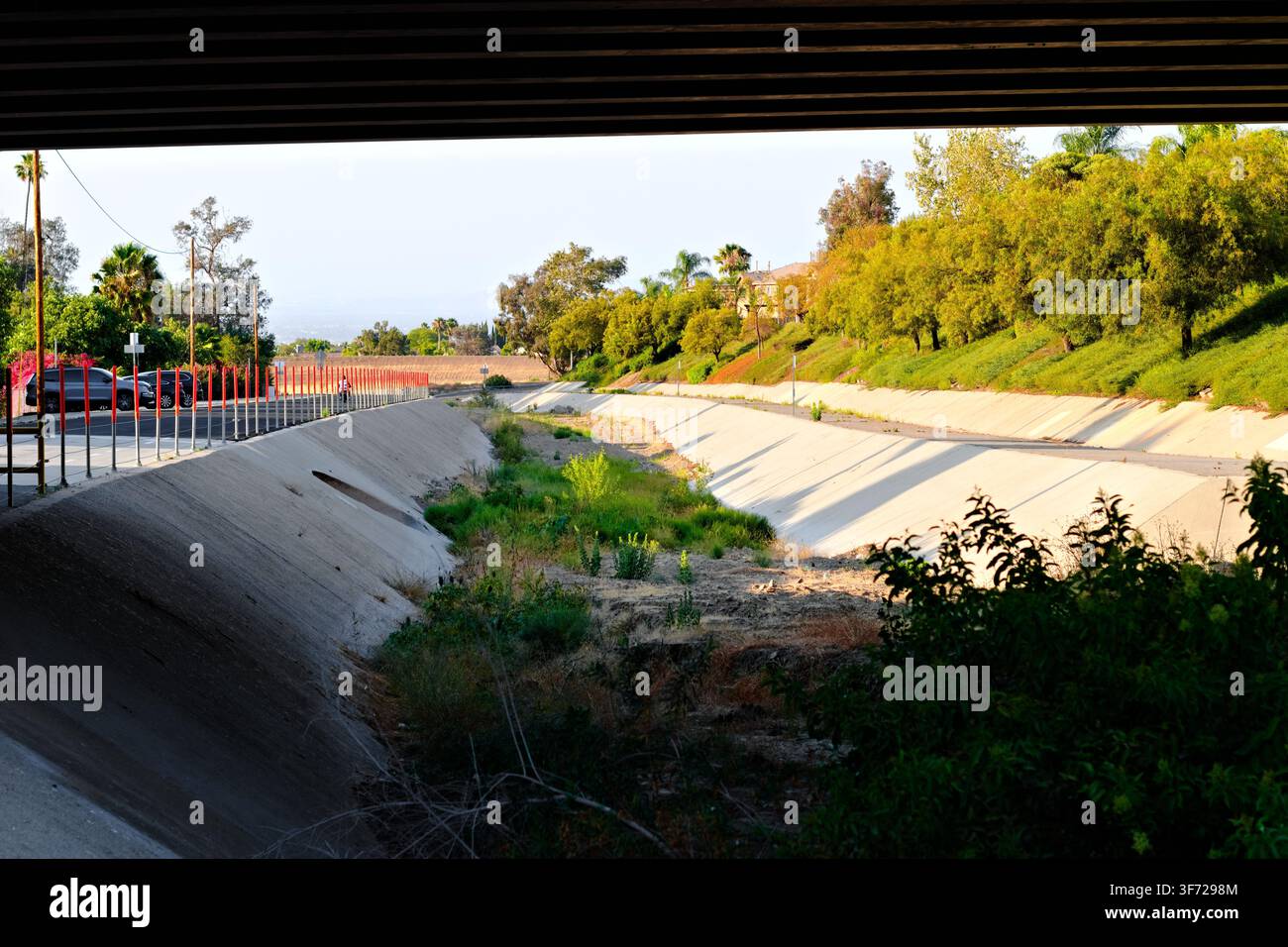 Ein betonierter Hochwasserschutzkanal verläuft unter einer Überführung neben Parkplätzen, Vegetation und landschaftlich gestalteten Hängen in Corona, Kalifornien. Stockfoto
