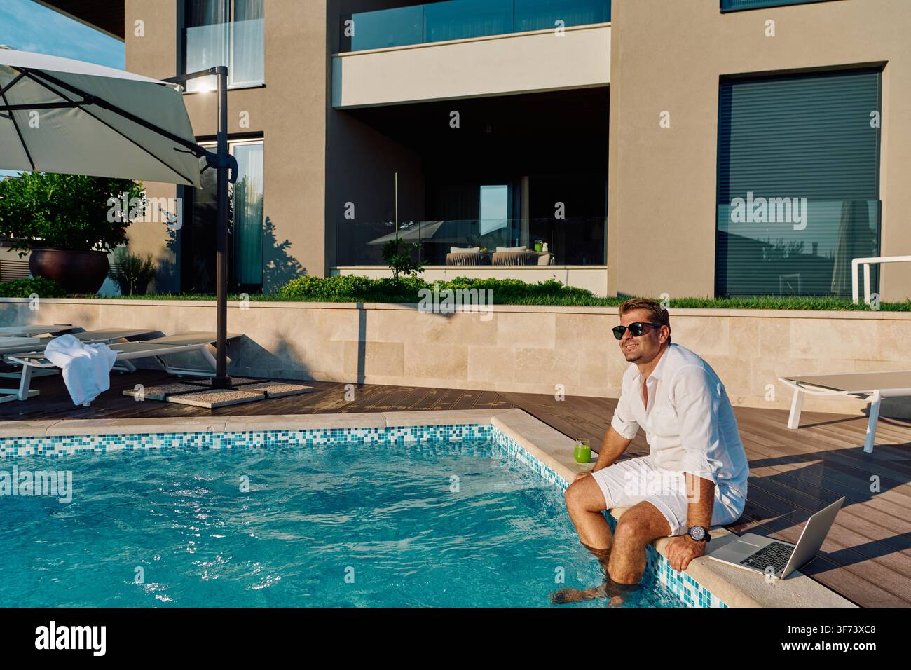 Ein Mann in Weiß sitzt am Rand eines Luxuspools mit seinem Laptop und einem Getränk, trägt eine Sonnenbrille und genießt einen entspannten Sommermorgen in einer modernen Villa, die Freizeit und Fernarbeit verbindet. Stockfoto