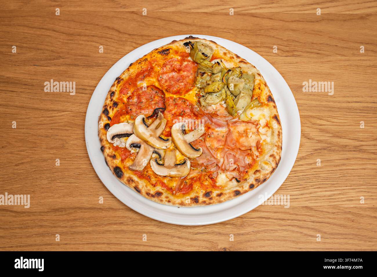 Vier-Jahreszeiten-Pizza mit Salami, Artischocken, Schinken und Pilzen auf einem weißen Teller über einem Holztisch. Stockfoto