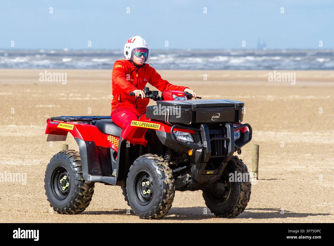 Ein RNLI-Lebensretter in roter Uniform, weißem Helm und Sonnenbrille fährt ein rotes Quad-Patrouillenfahrzeug. Southport, UK. Stockfoto