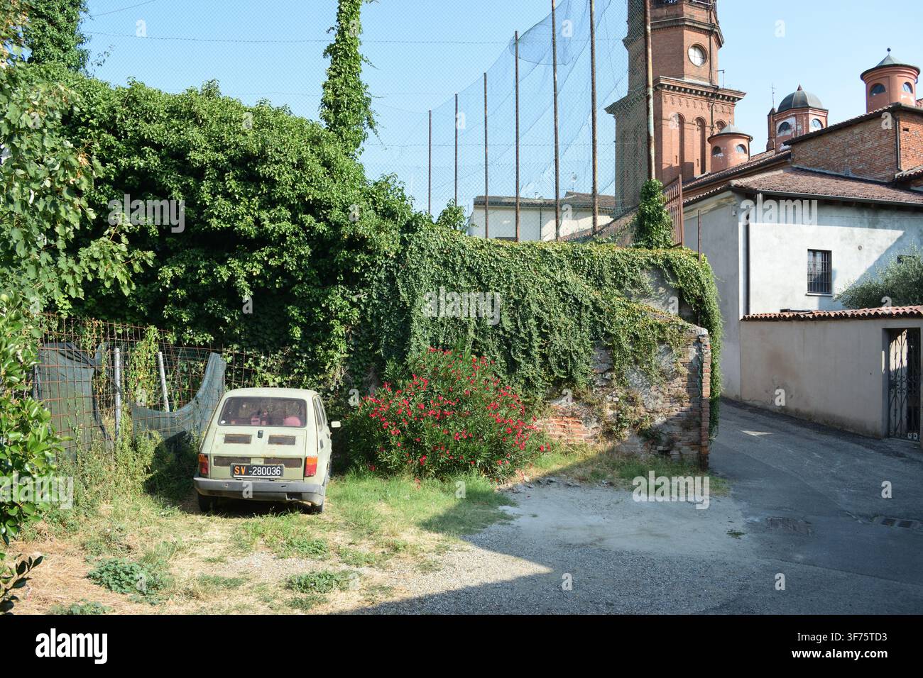 Klassischer hellgrüner Fiat 126, der auf einem grasbewachsenen Fleckchen geparkt ist, teilweise verdeckt von einem hell blühenden rosa Oleanderstrauch vor einem Drahtgitterzaun Stockfoto