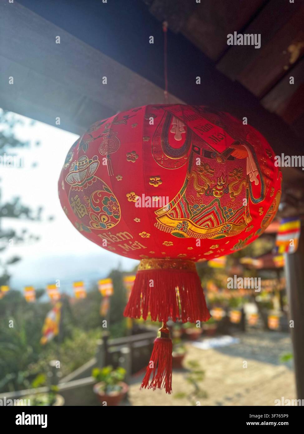 Traditionelle rote asiatische Laterne, die im Freien mit goldenen Verzierungen hängt. Helle dekorative Papiert Lampe mit aufwendigen orientalischen Mustern und einer Quaste. - Smartphone-aufgenommenes Stockfoto