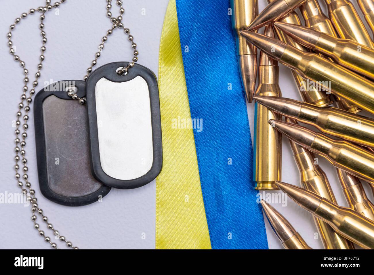 Metallene Hundemarken liegen neben Messinggewehrpatronen und einem blau-gelben Band auf einem hellen Hintergrund. Stockfoto
