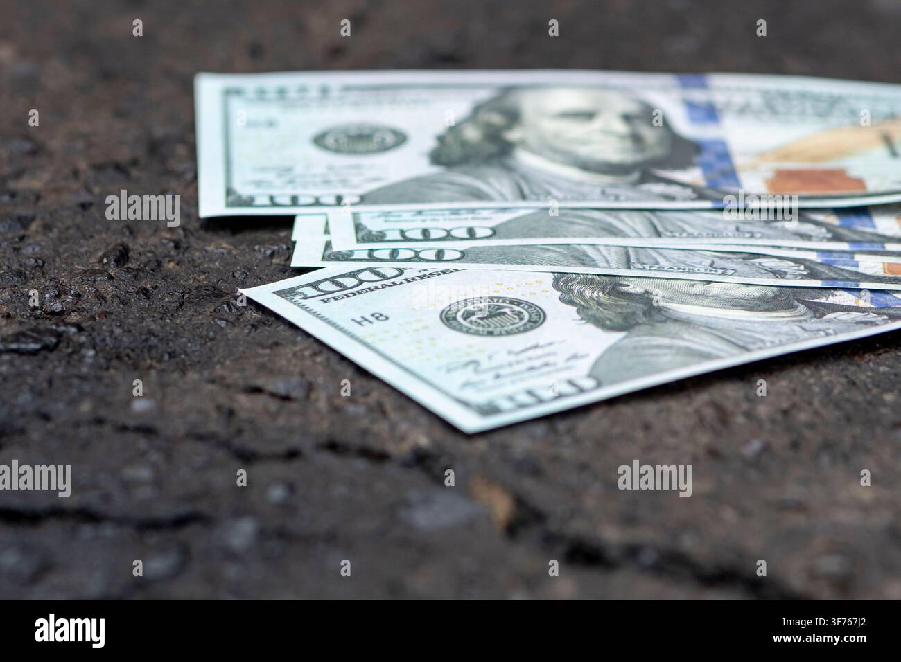 Mehrere Hundert-Dollar-Scheine sind über eine dunkle, raue Asphaltfläche verteilt. Stockfoto