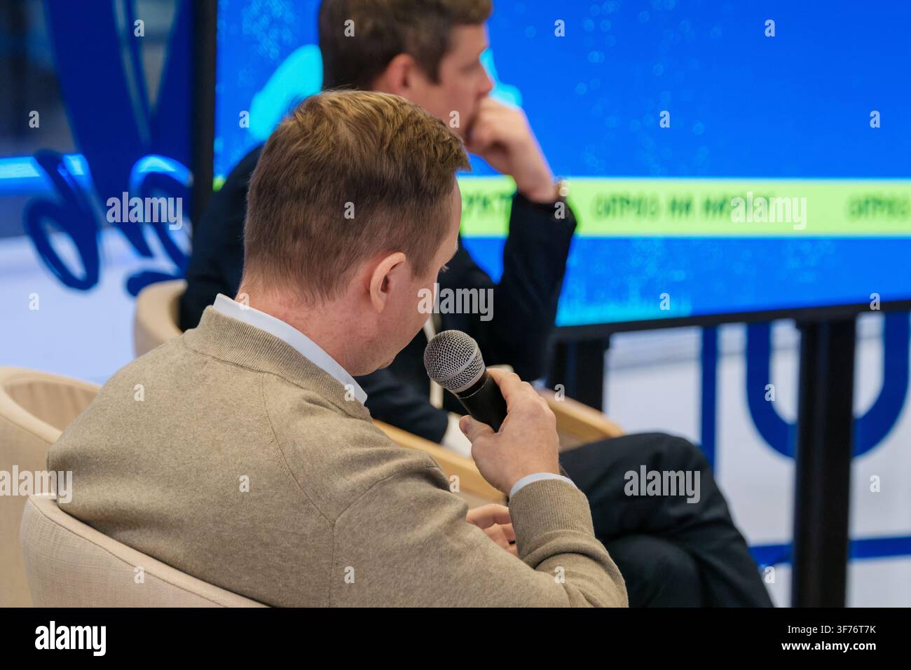 Sitzender männlicher Teilnehmer spricht in ein Handmikrofon, während ein anderer Teilnehmer hinter ihm zuhört. Stockfoto