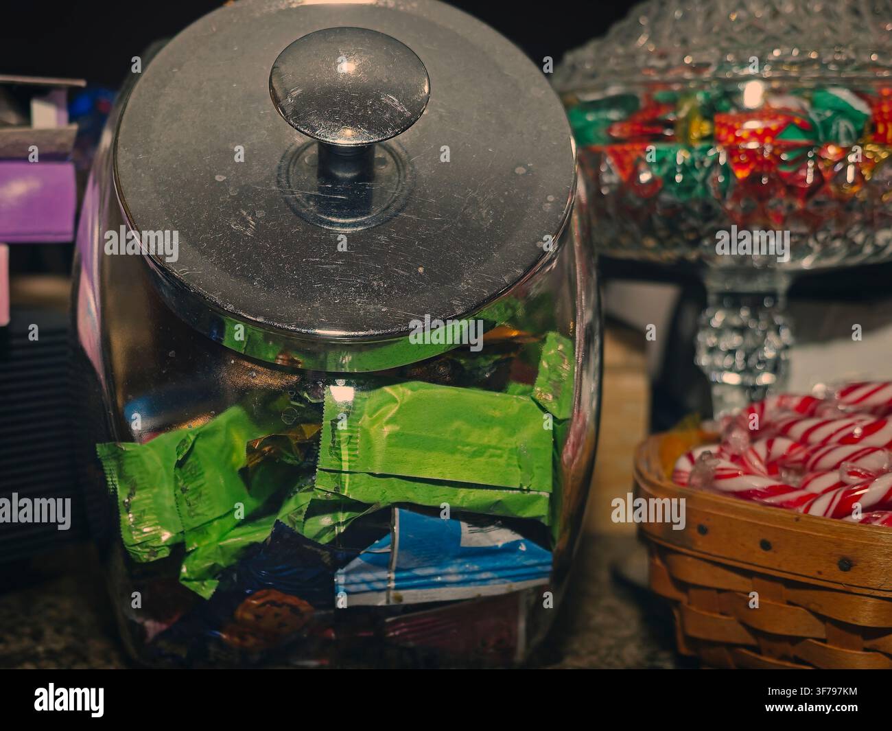Nahaufnahme eines großen Glasbonbongefäßes mit Metalldeckel, gefüllt mit bunten verpackten Bonbons, neben einem Korb mit Zuckerstangen Stockfoto