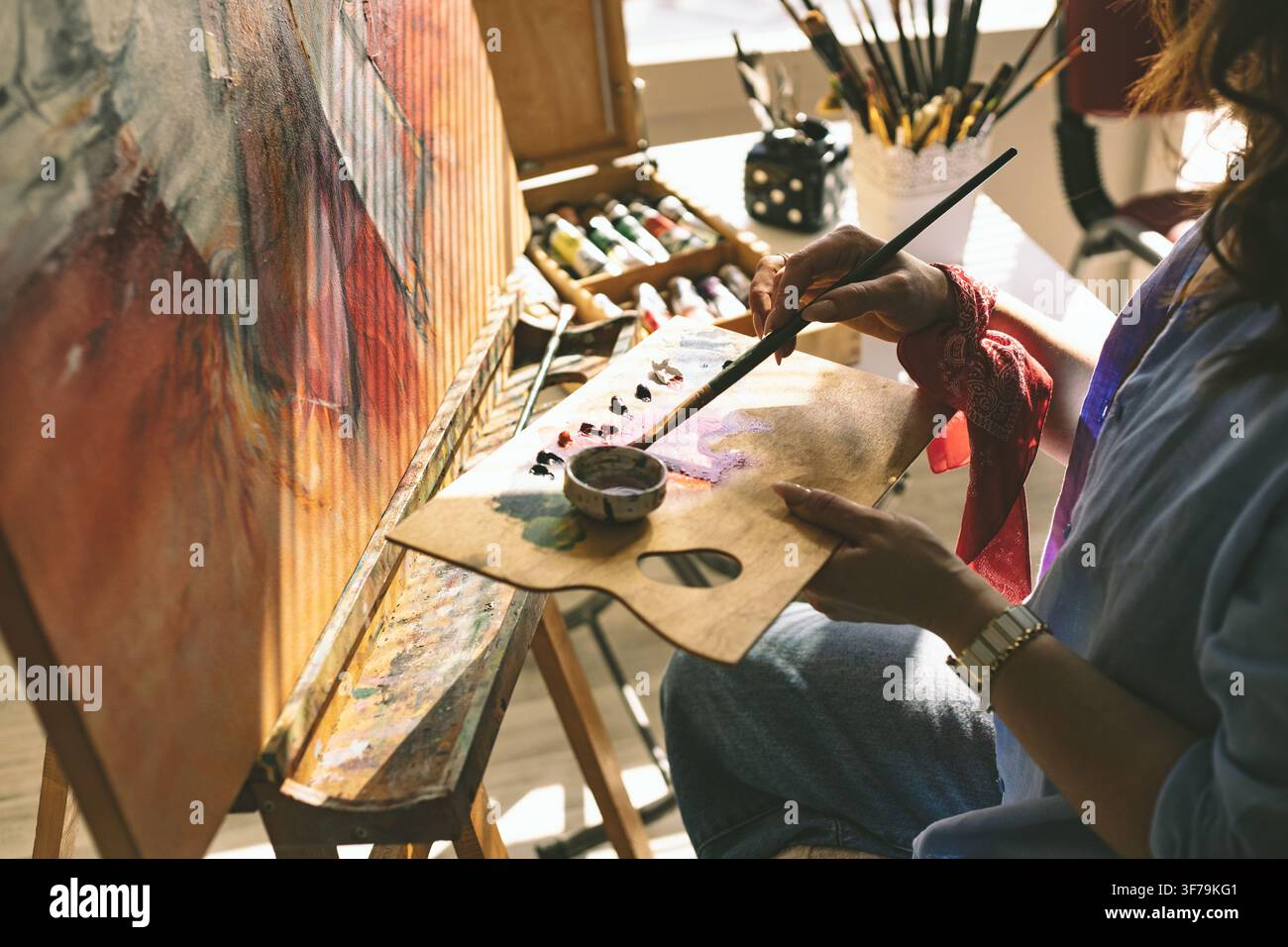 Nahaufnahme einer Künstlerin, die Farbe auf einer Palette mischt, während sie an einer Original-Leinwand in einem hell erleuchteten Atelier arbeitet. Stockfoto
