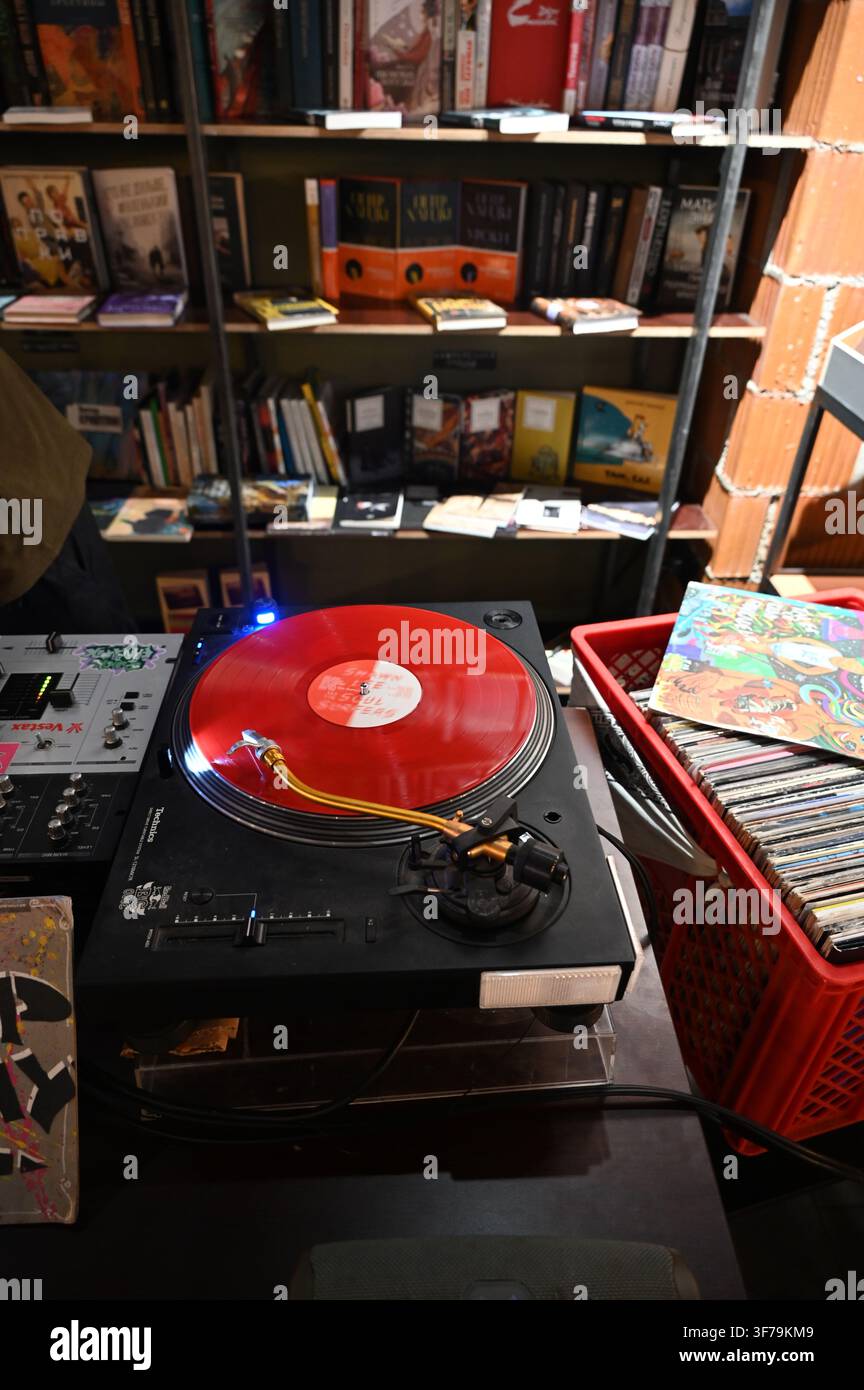 Ein horizontales Foto, das einen DJ-Teller zeigt, der eine lebhafte rote Vinylplatte abspielt, mit einer roten Kiste, die mit Platten und Hüllen gefüllt ist, daneben auf einem Tisch in einem Plattenladen positioniert. Stockfoto