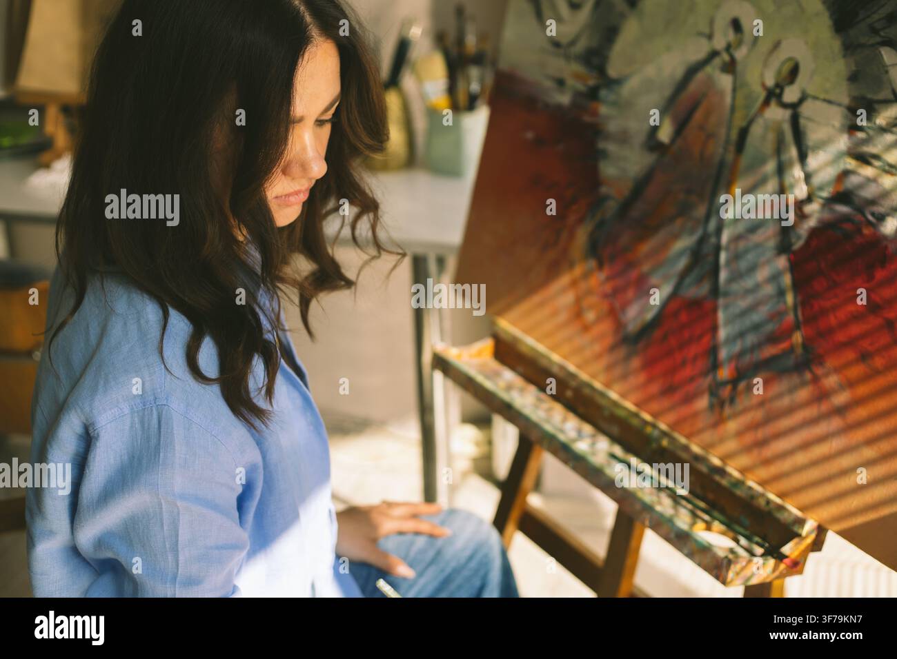 Nachdenkliche Künstlerin betrachtet ihr Originalgemälde in einem hellen Atelier, gefüllt mit natürlichem Sonnenlicht und kreativer Atmosphäre. Stockfoto