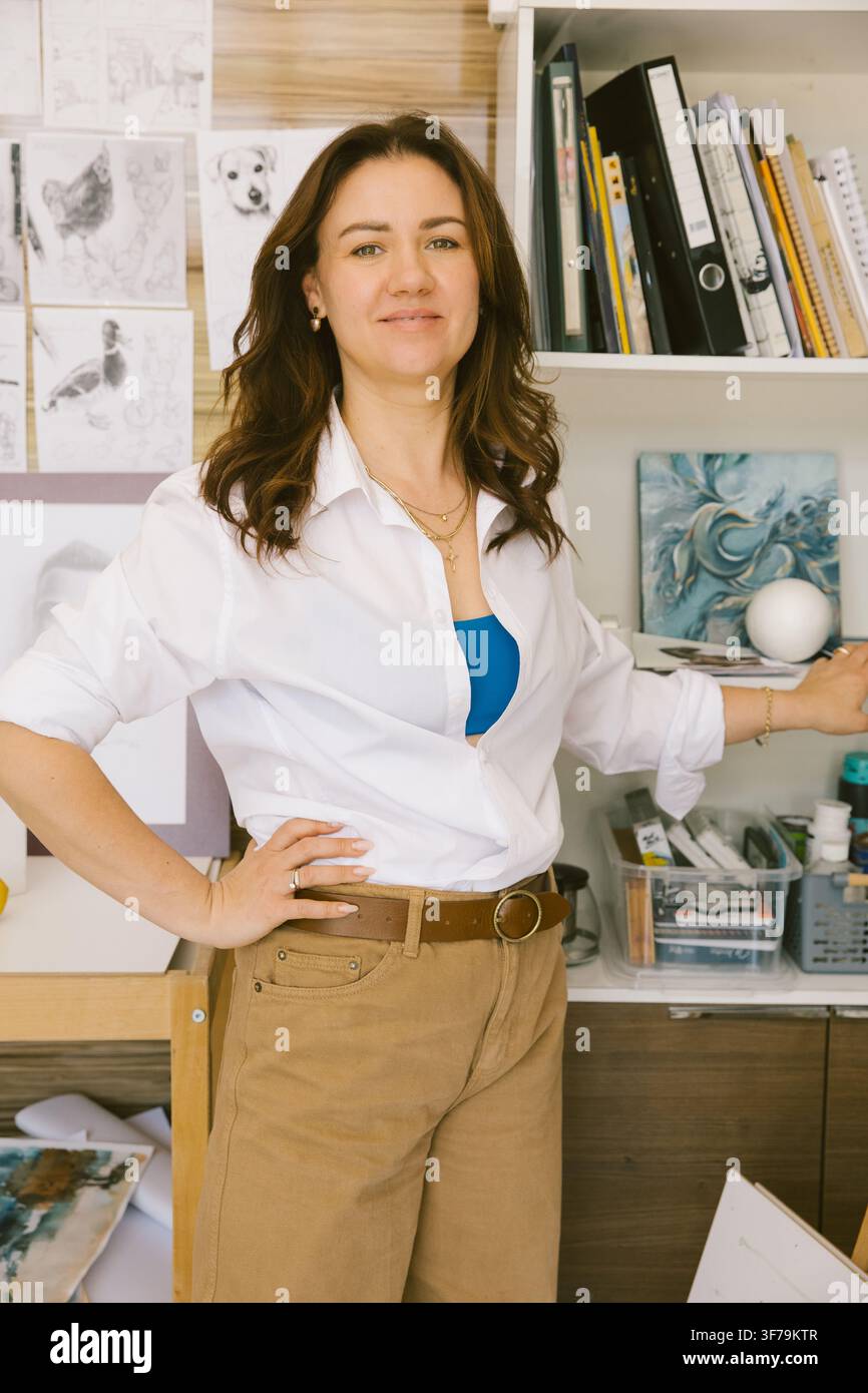 Vertikales Porträt einer lächelnden Künstlerin in ihrem kreativen Atelier, umgeben von Gemälden, Skizzen und Kunstmaterialien. Authentischer Lifestyle i Stockfoto