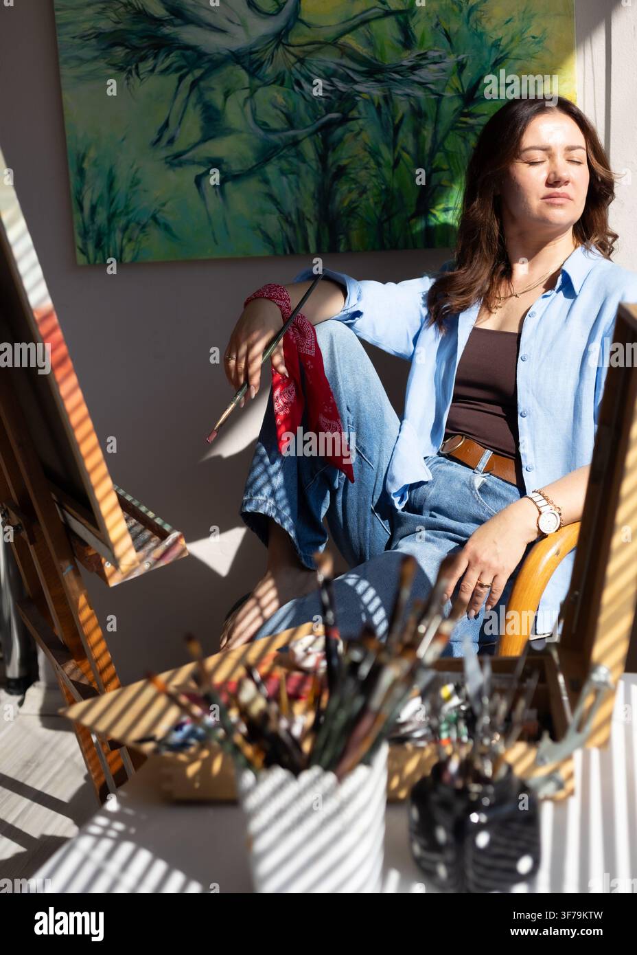 Porträt einer Künstlerin, die neben ihrer Staffelei in einem hellen Atelier mit warmem natürlichem Sonnenlicht entspannt. Stockfoto