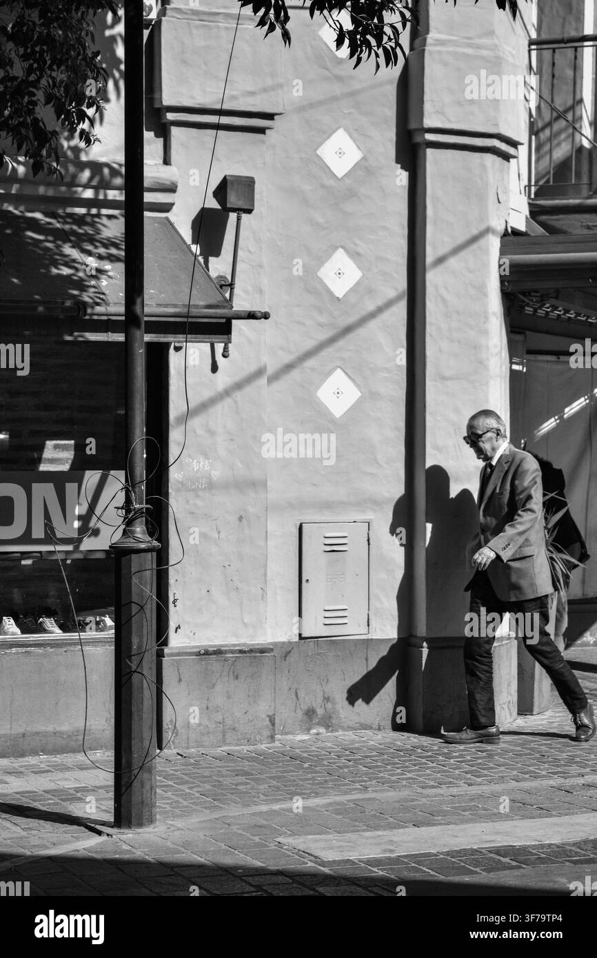 Senior mann in einer formellen Anzugjacke geht auf einem städtischen Bürgersteig neben starken architektonischen Schatten an einem sonnigen Tag. Stockfoto