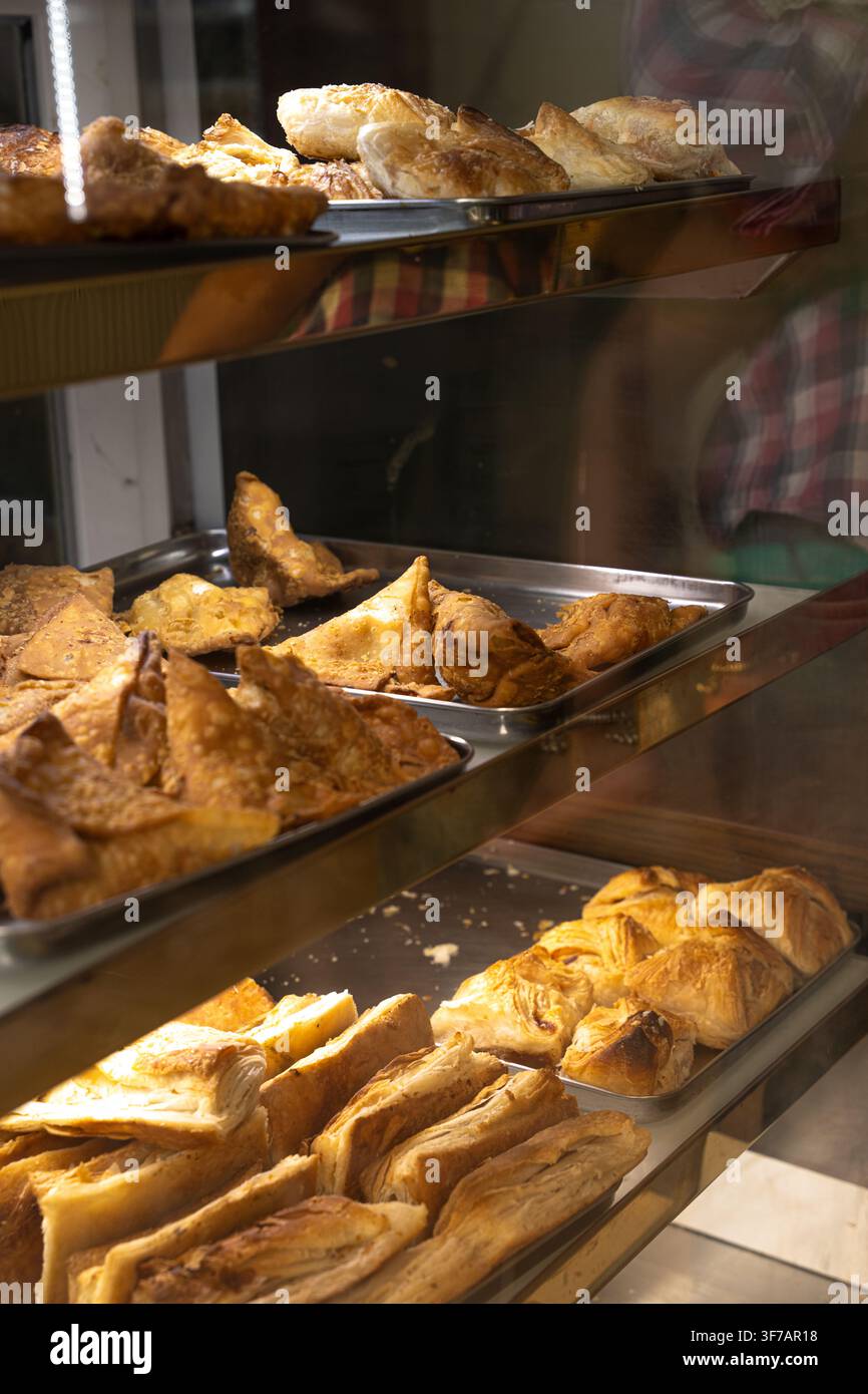 Goldene Samosas und blättrige Blätterteiggebäck sind auf mehreren Metalltabletts hinter reflektierendem Glas an einem Fertiggerichte-Schalter angeordnet. Stockfoto