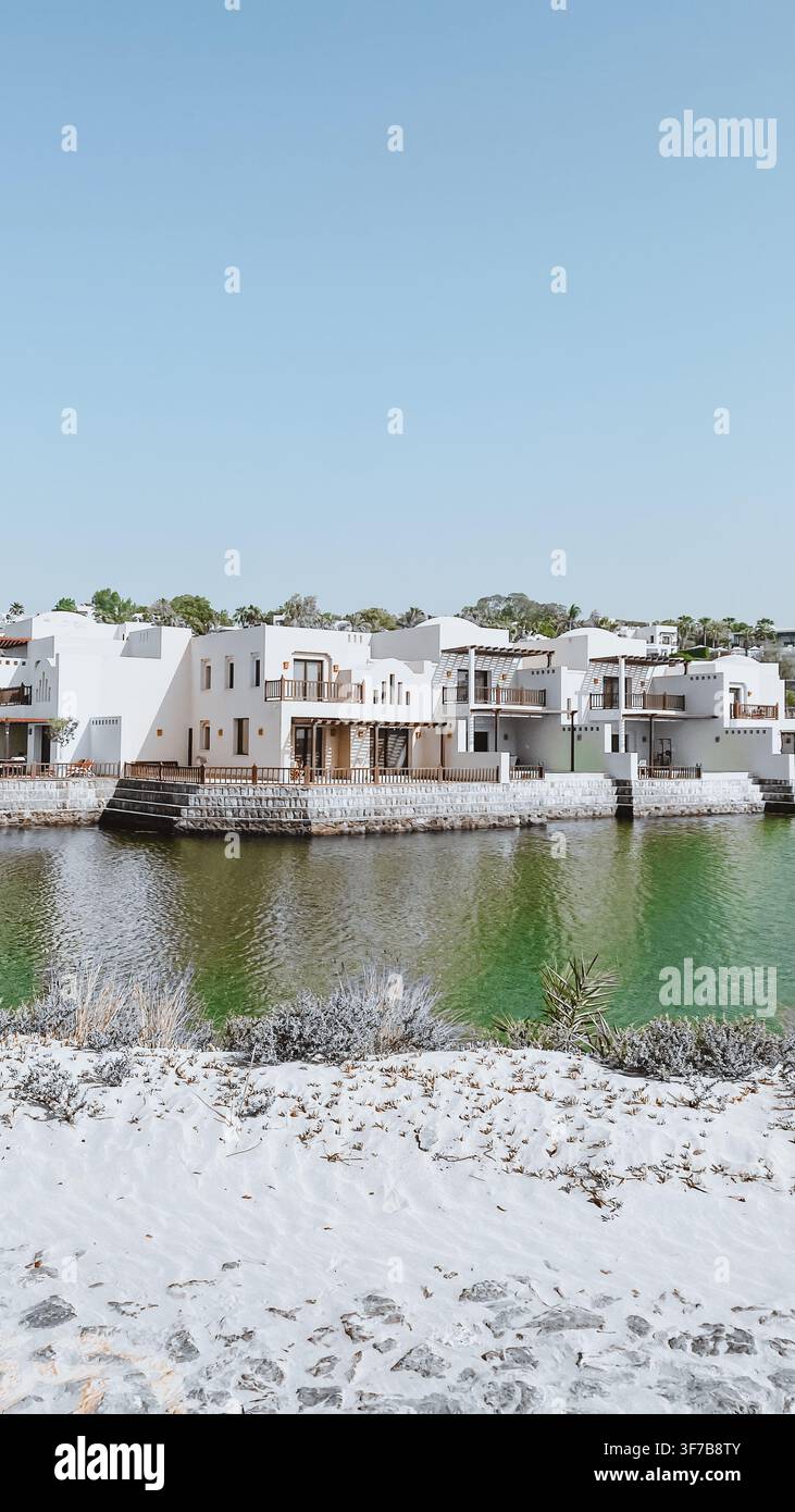 Weiße Ufervillen mit traditioneller nahöstlicher Architektur neben einer ruhigen Lagune unter einem klaren blauen Himmel. - Smartphone-aufgenommenes Stockfoto
