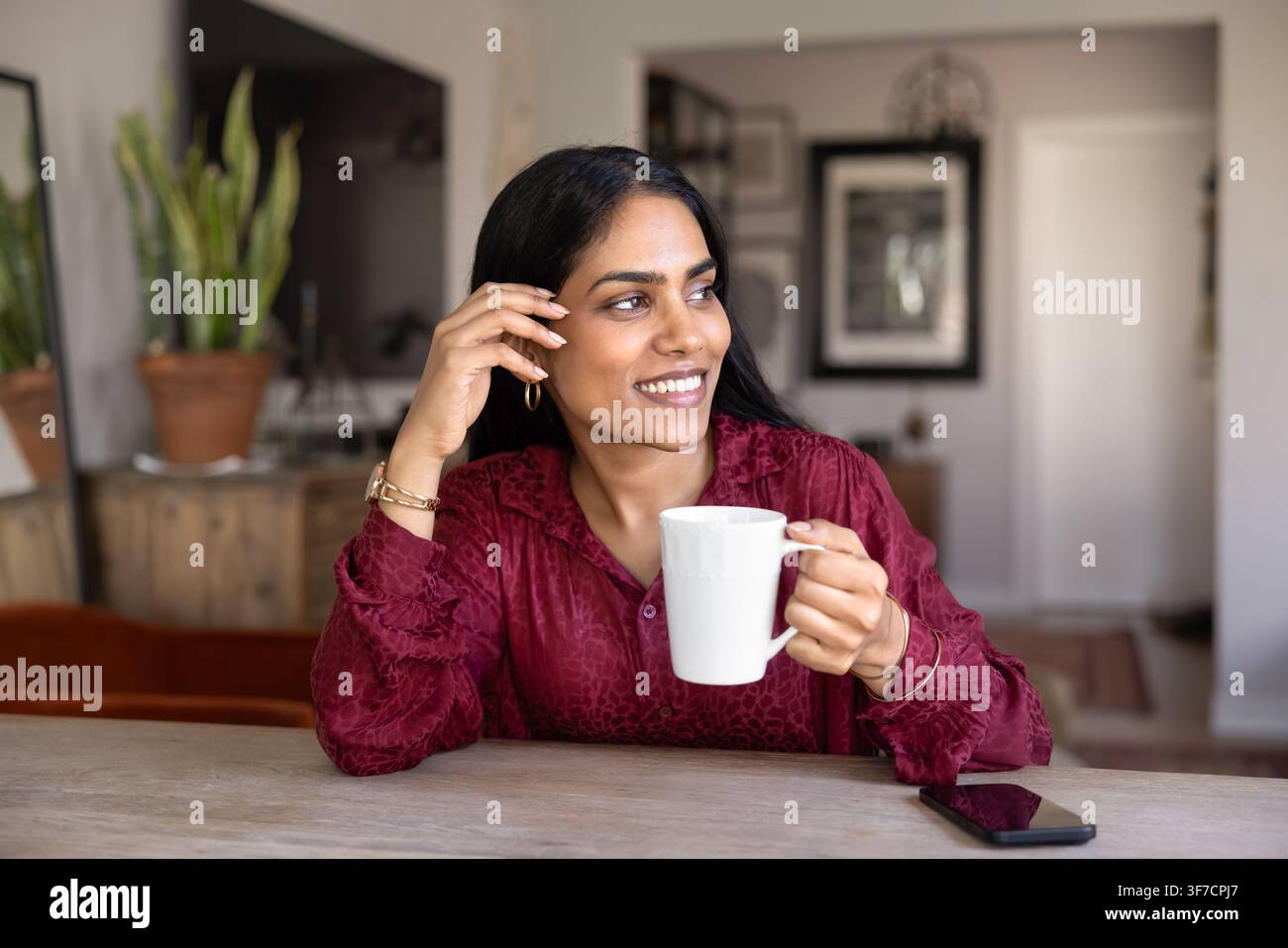 Lächelnde indische Frau sitzt an einem Tisch mit einer Tasse Stockfoto