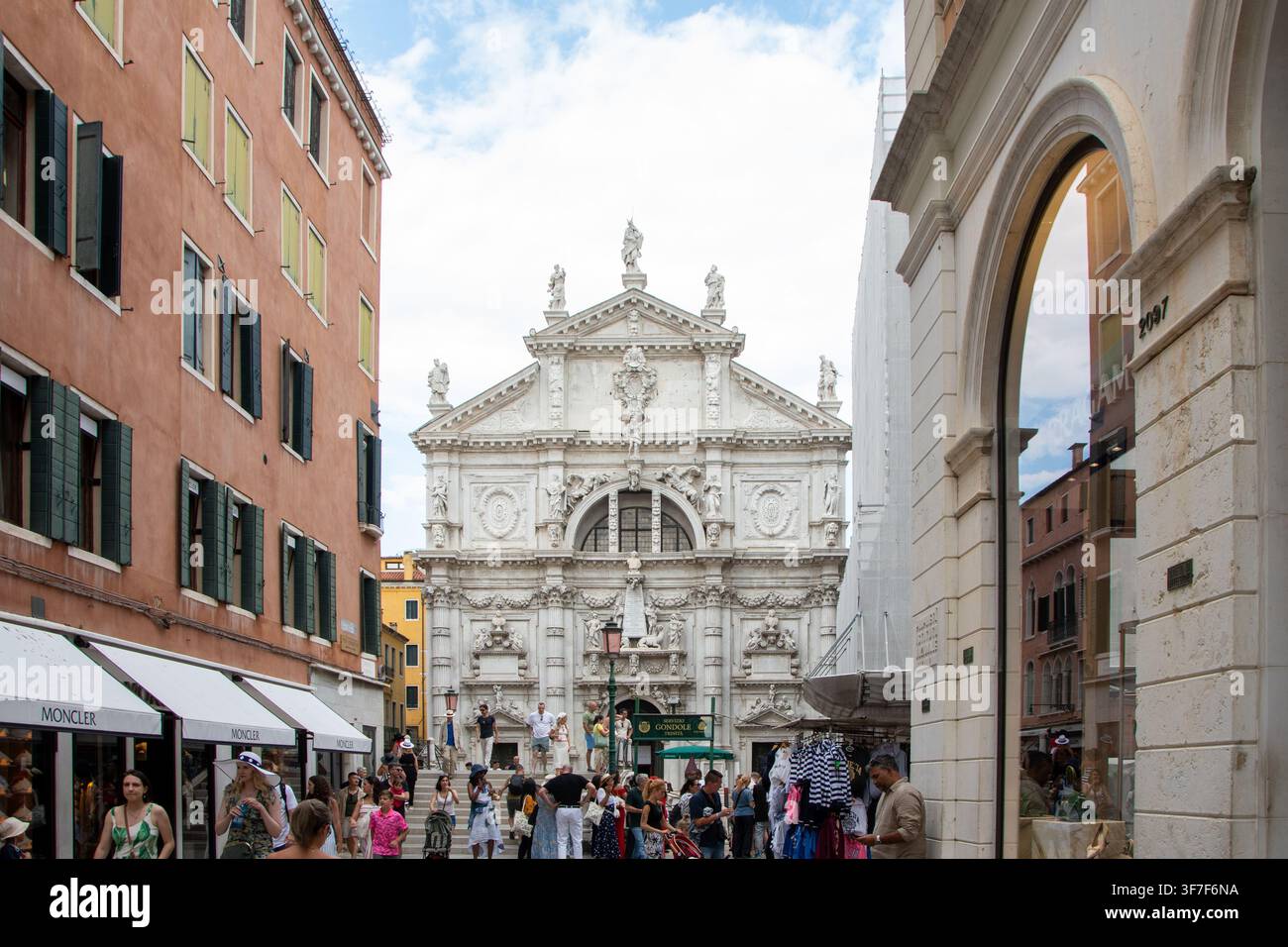 ITALIEN, VENEDIG, 11. Juli 2025: Eine Menschenmenge steht vor einer aufwendigen barocken Kirchenfassade zwischen hohen Gebäuden auf einer Stadtstraße. Stockfoto