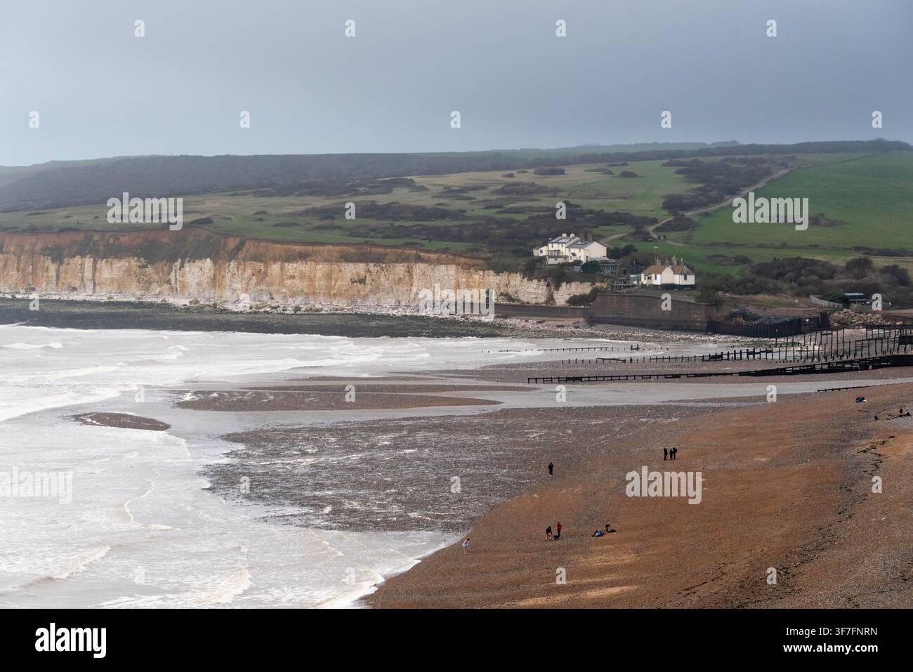 Besucher spazieren am Strand von Cuckmere Haven und genießen die Aussicht auf die historischen Coastguard-Cottages und die ikonischen Seven-Sisters-Klippen in East Sussex. Stockfoto