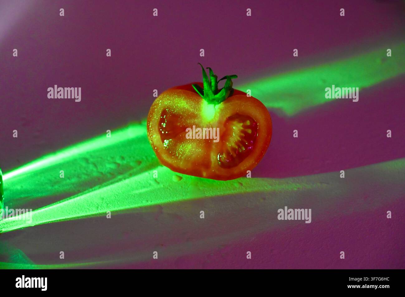 Nahaufnahme eines kommerziellen Bildes einer frischen reifen roten Tomatenhälfte mit einem grünen Stiel, zentriert unter neongrünen Lichtstrahlen vor einem dunklen violetten und rosa texturierten Hintergrund. Geeignet für konzeptionelle Foodfotografie, modernes kulinarisches Design und kreative Werbung. Stockfoto