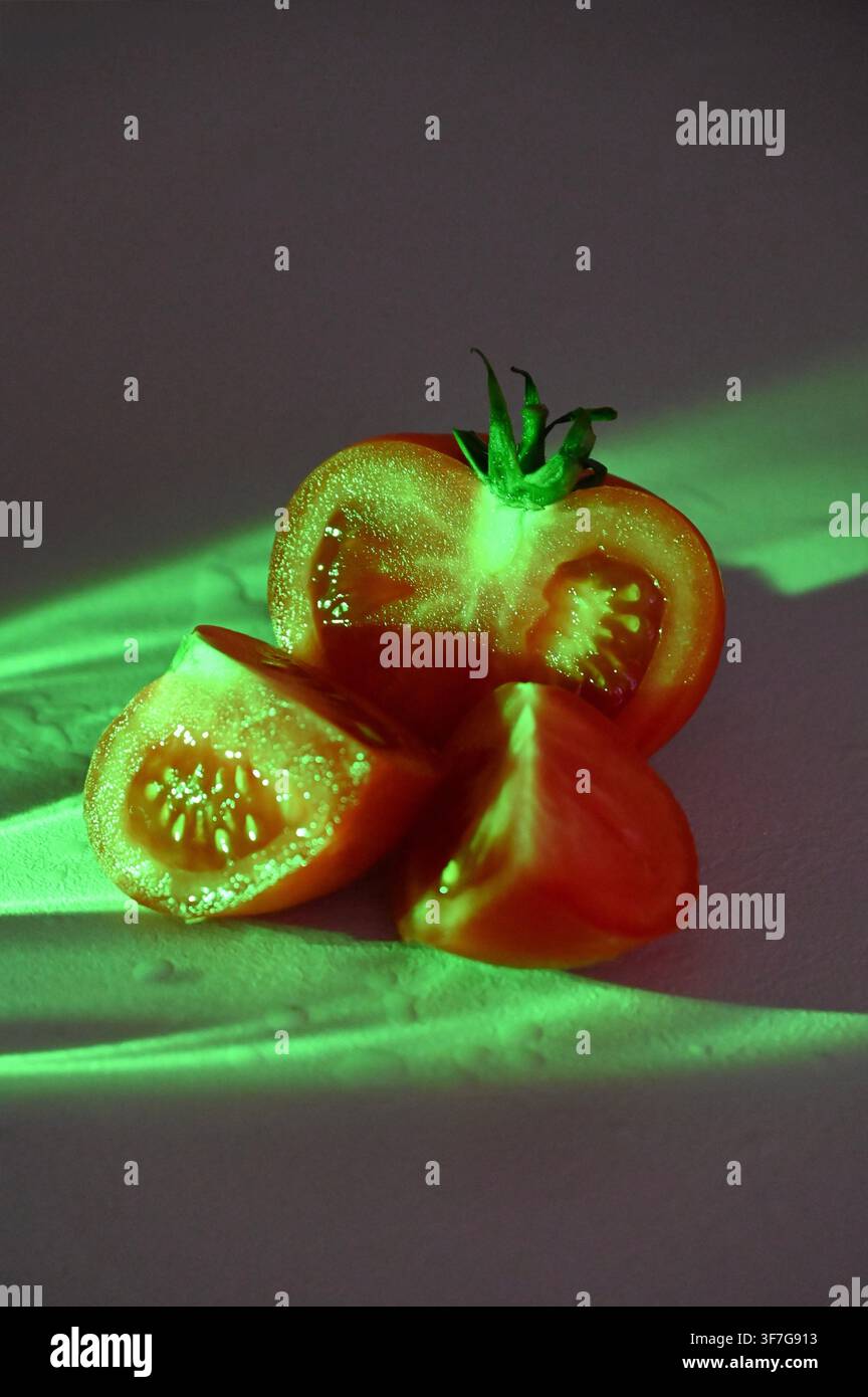 Nahaufnahme einer kommerziellen Aufnahme einer frischen reifen roten Tomate, die in Hälften und Viertel geschnitten ist und einen grünen Stiel hat, beleuchtet von neongrünen Lichtstrahlen vor einem dunkelvioletten und rosa texturierten Hintergrund. Geeignet für konzeptionelle Food-Fotografie, modernes kulinarisches Design und kreative Werbung. Stockfoto
