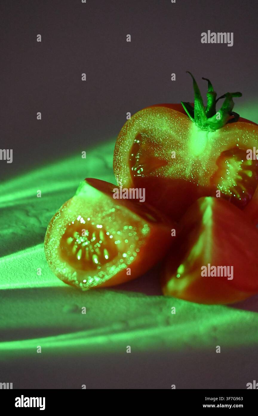 Nahaufnahme einer kommerziellen Aufnahme einer frischen reifen roten Tomate, die in Hälften und Viertel geschnitten ist und einen grünen Stiel hat, beleuchtet von neongrünen Lichtstrahlen vor einem dunkelvioletten und rosa texturierten Hintergrund. Geeignet für konzeptionelle Food-Fotografie, modernes kulinarisches Design und kreative Werbung. Stockfoto