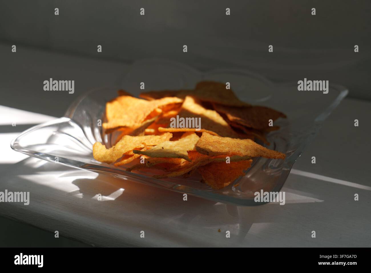Dreieckige Tortilla-Chips füllen eine klare Glasschale, werden von starkem Sonnenlicht beleuchtet, das kontrastierende Schatten wirft und die knusprige Textur des Snacks hervorhebt Stockfoto