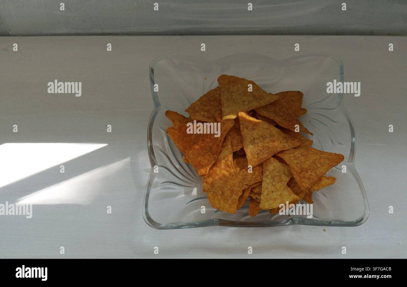 Nacho-Tortilla-Chips füllen eine Glasschale und bieten einen knusprigen und salzigen Snack auf einer weißen Oberfläche, wobei natürliches Fensterlicht einen Schatten wirft Stockfoto