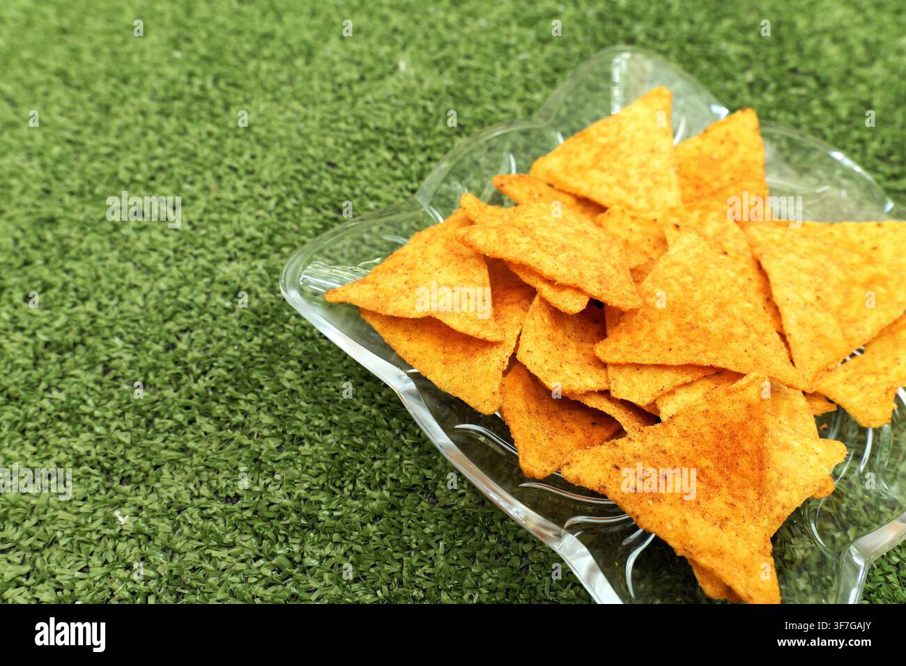 Nacho-Chips mit Gewürzfüllung in einer klaren Glasschale, die eine herzhafte Snack-Präsentation gegen einen lebendigen grünen Kunstrasenhintergrund bildet, perfekt fo Stockfoto