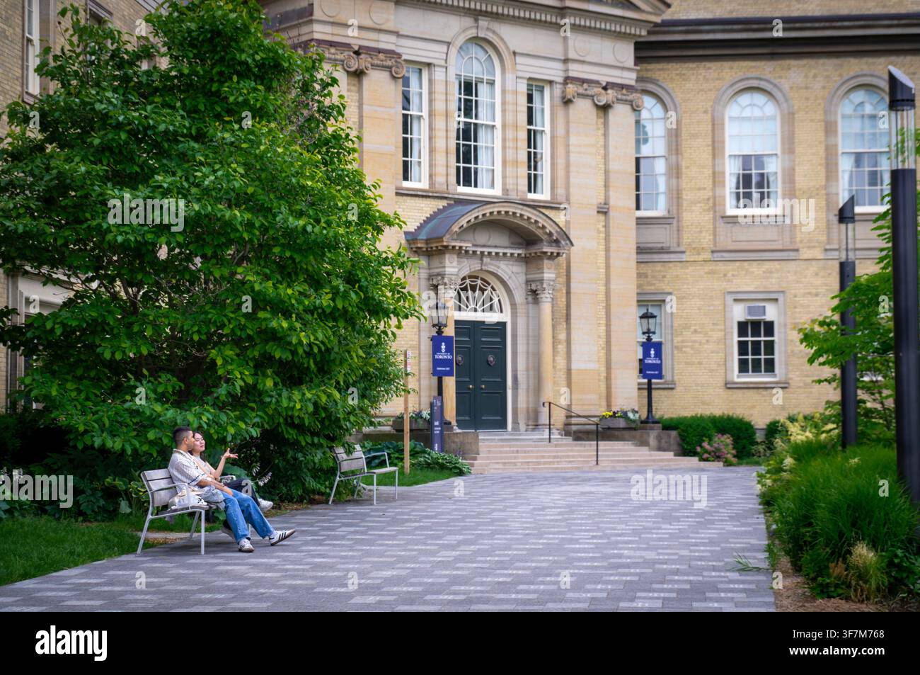 Toronto, Ontario, Kanada – 06 21 2025: Ein junger Mann und eine Frau unterhalten sich auf einer Campusbank vor einem klassischen sandsteinfarbenen akademischen Gebäude mit gewölbten Fenstern Stockfoto