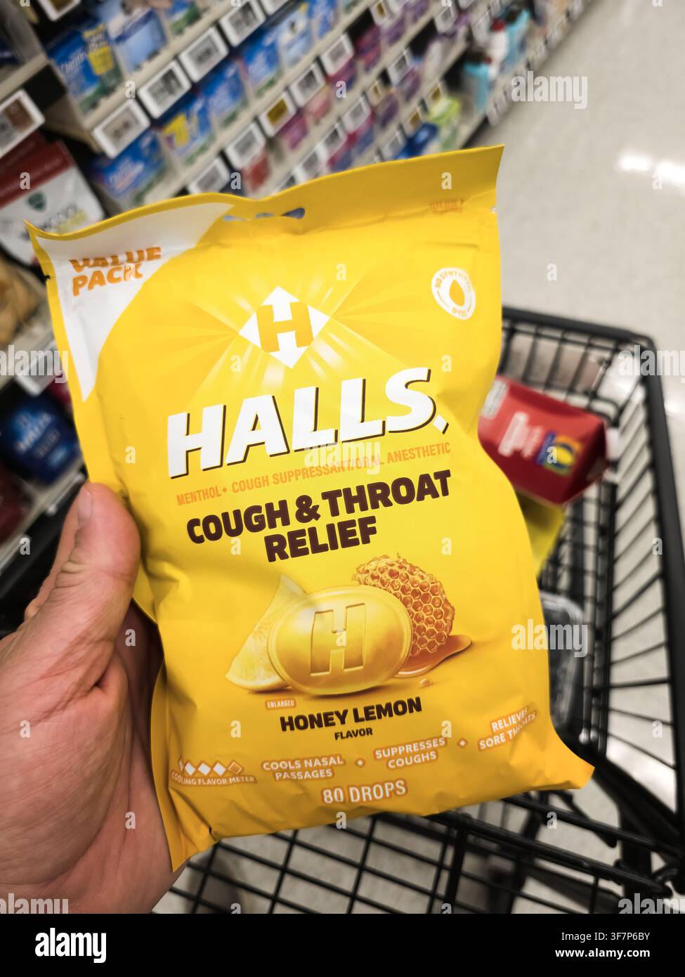 ADDISSON, TX, USA - 21. JULI 2026: Eine Hand, die eine Tüte Halls Honey Lemon Cough and Throat Relief-Tropfen in einem Einkaufswagen hält, vor einem Hintergrund o Stockfoto