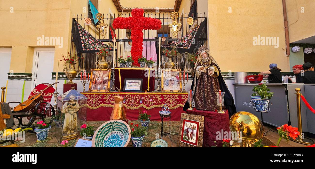 Ein rotes, mit Blumen bedecktes Mai-Kreuz erhebt sich über eine aufwendige Hofdarstellung mit einer bekleideten religiösen Figur, bestickten Schals und gerahmten Andachtsbildern Stockfoto