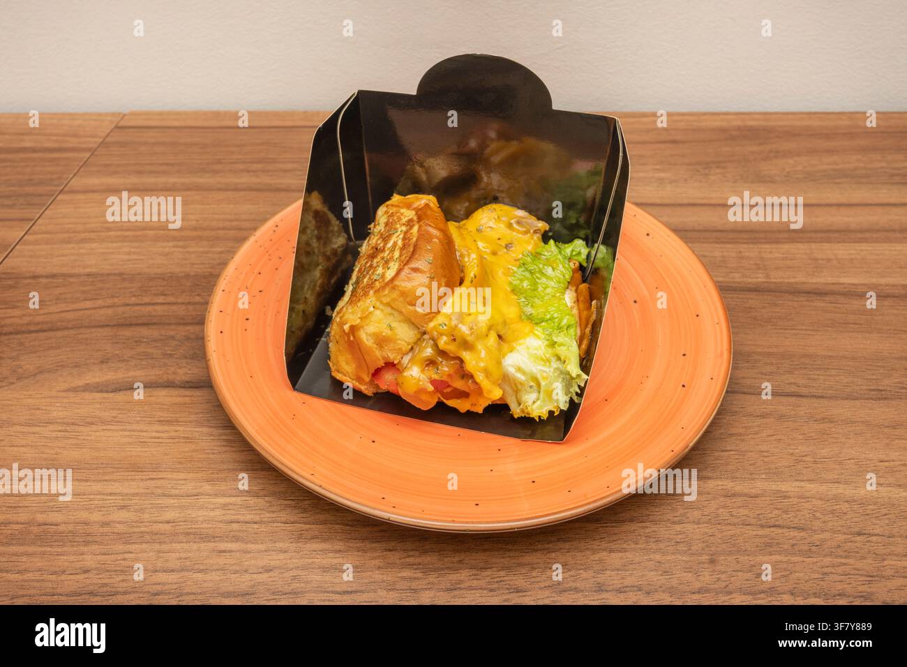Käsesandwich mit Salat und Tomate in schwarzem Papierhalter, serviert auf einem orangefarbenen Teller. Stockfoto
